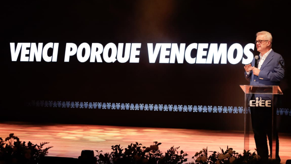 Humberto Casagrande, CEO do CIEE, aparece no palco durante a cerimônia, posicionado ao lado de um púlpito transparente com o logo do CIEE. Ele usa terno azul e fala ao público enquanto, ao fundo, um telão exibe a frase “Venci porque vencemos
