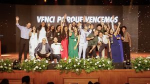 Grupo de formandos do projeto Somos CIEE posa no palco durante a cerimônia de formatura. Eles estão sorrindo, alguns com as mãos erguidas em comemoração. Ao fundo, um telão exibe a frase “Venci porque vencemos